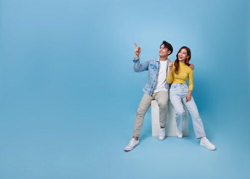 Happy Asian couple sitting on white cube chair and presenting or showing hand pointing finger with for product isolated on blue studio copy space background.