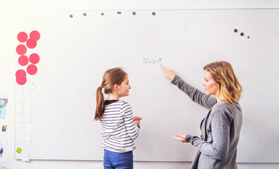 Hardworking teacher looking at young schoolgirl in classroom. Girl student writing on whiteboard.