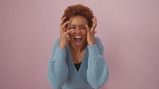 Frustrated african american woman in sweater, standing enraged, arms raised, yelling with wild, manic expression before pink backdrop