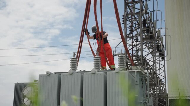 A construction working is tying the ropes hanging from a crane onto the corners of a transformer so it can be carried. Sunny and cloudy day