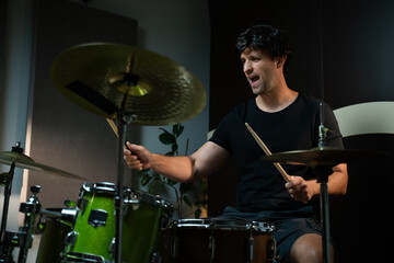 A man dressed in a black T-shirt and shorts sits at a drum kit in a home studio. He plays the drums with wooden sticks