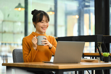 Pretty young female freelancer holding coffee cup and reading email on her laptop