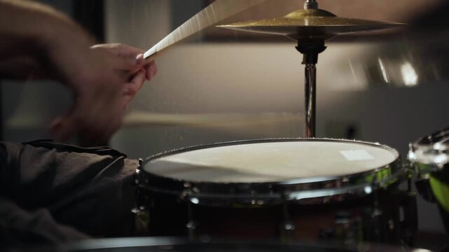A close-up shot of a drummer's hands striking a snare drum with drumsticks, creating a shower of water droplets. The drum is in focus, and the background is blurred. The drummer is wearing a dark