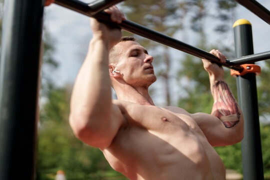 An athletic man with visible muscles is working out on pull-up bars in a sunny outdoor park, illustrating strength and fitness amidst nature.