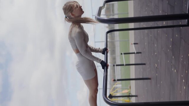 A focused young woman in athletic wear using parallel bars at a park, showcasing determination and fitness under a cloudy sky. Young Woman Training on Outdoor Parallel Bars Against Sky. Vertical video
