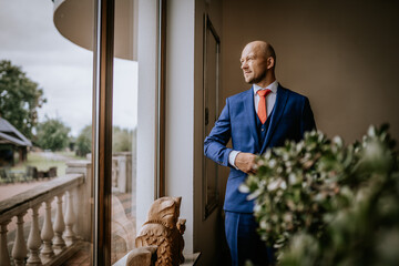Cesis, Latvia - July 12, 2024 - A man in a blue suit and red tie stands by a large window, looking...