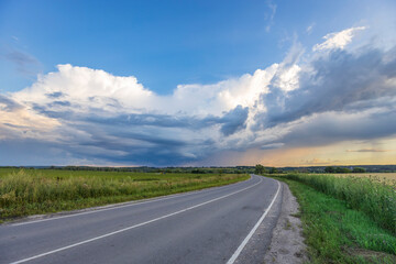 A paved road curves through a green field, disappearing into the distance beneath a dramatic, cloudy sky.