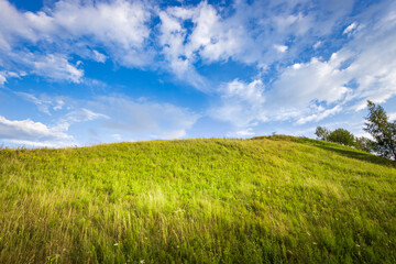 Vibrant green grass covers the gentle slope, while the sky shines bright with fluffy clouds,...