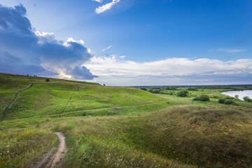 Fototapeta premium Rolling green hills gently slope towards a tranquil river, as dramatic clouds loom overhead, creating a picturesque evening landscape.