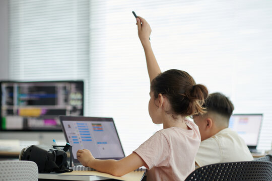 Students participating in computer class, one raising hand while answering a question Teacher providing guidance on programming tasks Classroom filled with computers and technological equipment