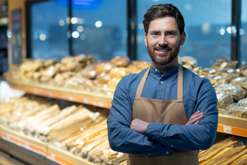 Confident baker standing with arms crossed in bakery shop surrounded by fresh bread. Person showcasing pride in craftsmanship and dedication to baking. Background displays variety of breads