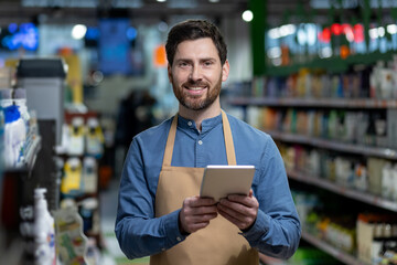 Smiling supermarket worker using digital tablet for inventory management in store aisle. Employee wearing apron holding tablet, performing inventory management tasks. Modern retail technology