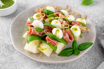 Appetizer with melon, mozzarella, prosciutto ham and fresh basil on stick with pesto sauce on a stone background. Finger food. Summer salad.