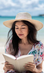 A woman with long dark hair wearing a straw hat and a floral blouse, reading a book on the beach. The ocean waves are visible in the background, creating a serene atmosphere.