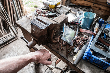 Farm worker seen turning a metal vice on a workbench within a barn. Various cables, nails can be seen on the grimy workbench.