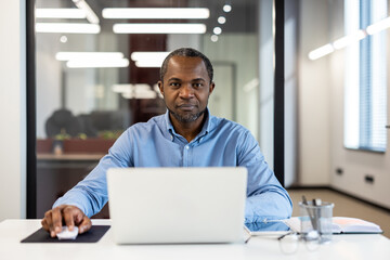 Confident businessman working on laptop in modern office. Focused professional using computer and mouse, performing work duties. Bright, modern office background.