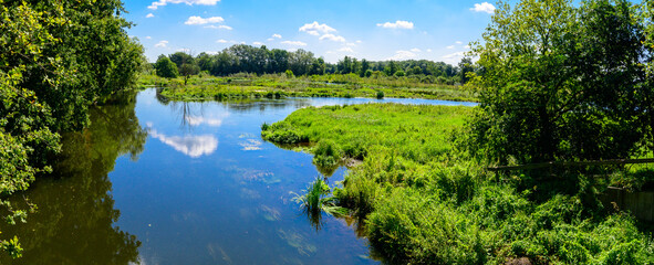 Panorama photo in the middle of summer with a view of a river/lake surrounded by green forests reflecting in the water. With blue whole with white clouds.