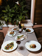 beautiful wooden table lies a gray tablecloth with delicious dishes and two glass goblets of wine. Next to the table stands a green plant in a cozy and nice restaurant