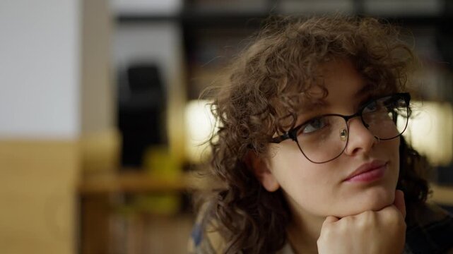 Close-up of a pensive girl student in glasses with curly hair looks to the side while thinking about her classes at the university