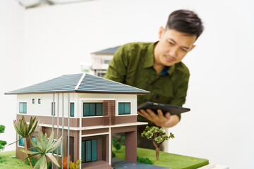 Asian businessman works alone at his desk, surrounded by a laptop, paperwork, tablet, and calculator, focusing on home and land mortgage agreements in a modern house model.