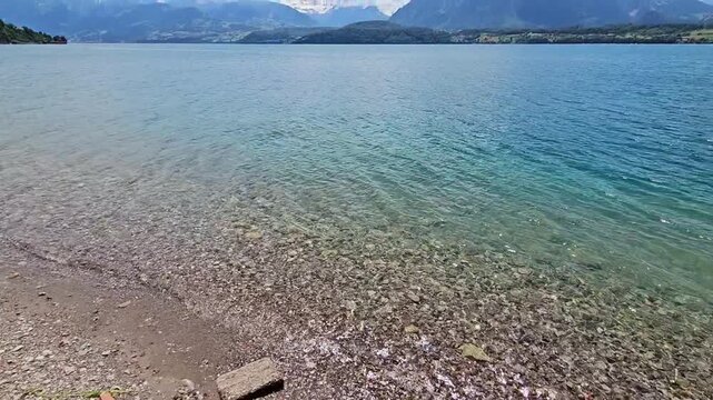 Static shot of shore of lake with clear water and mountains in background in Switzerland.
