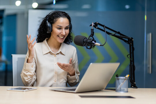 Happy woman in headphones using laptop and microphone for podcast in modern office. Smiling, engaging with audience, and conveying excitement. Professional setting with tablet and phone on desk.