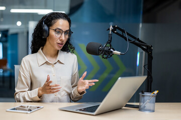 Confident woman wearing headphones recording podcast with laptop and microphone in modern office....