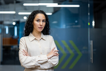 Confident businesswoman standing with arms crossed in modern office. Professional attire, modern...
