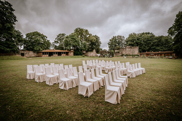Cesis, Latvia - July 12, 2024 -  Rows of white chairs are set up outdoors on a green lawn, ready for an event, with trees and historic ruins in the background under a cloudy sky.
