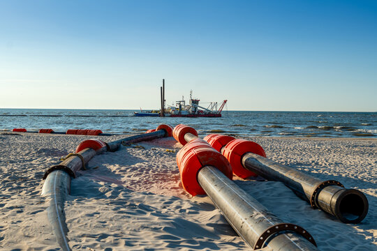 Marine works on dredging the bottom of the Baltic Sea. Two dredger ships with crew at work on sunny, summer day. Pipes lying on the beach in the foreground