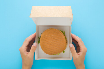 Young adult woman hands holding opened carton box with fresh burger on light blue table background....