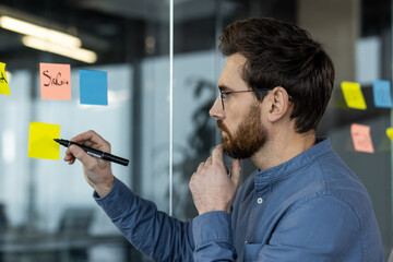 Man brainstorming with sticky notes on glass wall in modern office