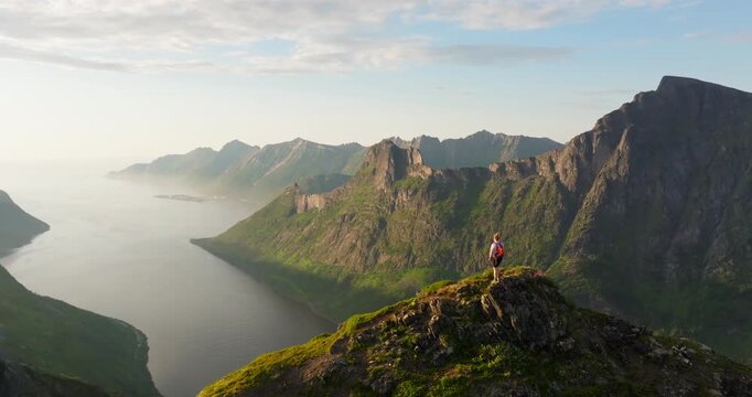Female on arctic Norway mountain top look at fjord in sunset glow; drone reveal
