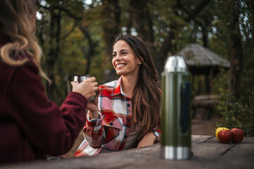 Happy young women friends taking a break from hing in sunny field. Two women relaxing in nature, they love autumn and each other, drinking hot drink