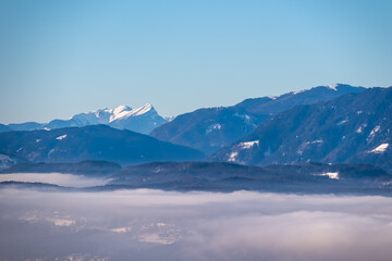 Panoramic view of snow capped mountain peak Latschur in winter seen from tower Pyramidenkogel, Carinthia, Austria. Fog covered alpine valley surrounded by majestic Austrian Alps. Mystical atmosphere