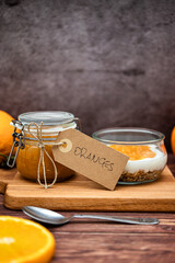 Glass jar full of muesli and yogurt with homemade orange jam, delicious snack with cheesecake and orange jam on wooden cutting board, detail with vintage cardboard tag, vertical image for social