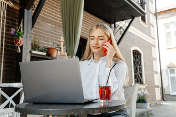 young beautiful blond caucasian woman sitting at table in outdoor terrace of cafe in summer day with notebook, businesswoman working, freelancer talking by phone and smiling