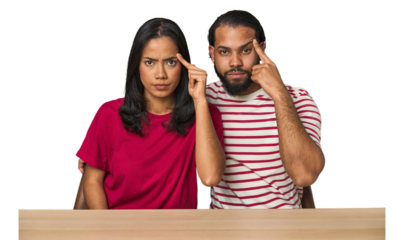 Seated young Latino couple at table pointing temple with finger, thinking, focused on a task.