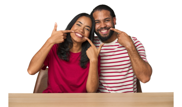 Seated young Latino couple at table smiles, pointing fingers at mouth.