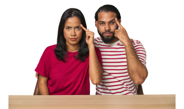 Seated young Latino couple at table pointing temple with finger, thinking, focused on a task.