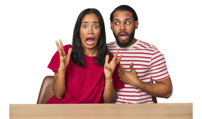 Seated young Latino couple at table screaming to the sky, looking up, frustrated.