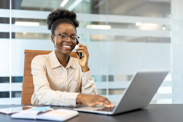 Young African American businesswoman working in office. Employee on phone and laptop smiling, wearing glasses and white shirt at desk. Professional workplace environment