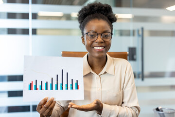 Young African American businesswoman at workplace holding data chart. Confident employee presenting statistical graph in modern office setting while smiling at camera.