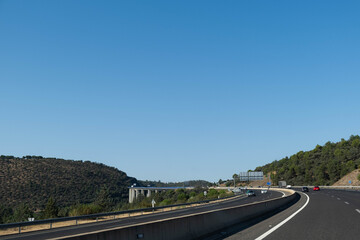 Panoramic view of a huge curved highway with a large bridge in the background and cars driving in Spain. The day is clear and sunny and the sky is blue.