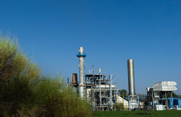 Exterior view of an industrial paper factory surrounded by green plants. It has two chimneys....