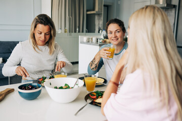 Three middle-aged women are talking over breakfast together in a cozy apartment.