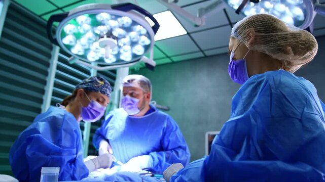 Scrub nurse stands watching the doctors operating the patient. Low angle view. Female surgeon turns to table with tools taking one.