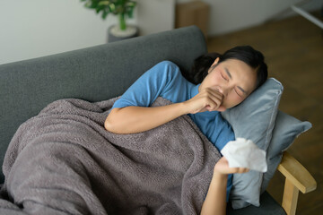Young Woman Resting on Sofa with Blanket, Holding Tissue and Coughing, Feeling Unwell at Home
