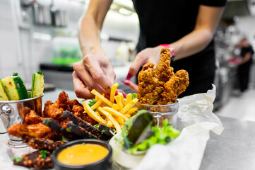 Close-up of hands preparing a food platter with fried chicken, fries, and fresh vegetables in a modern kitchen setting. vibrant colors and fresh ingredients highlight a delicious and appetizing meal.