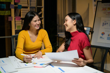 Obraz premium Two Young Women Collaborating on Business Project in Modern Office Setting with Charts and Graphs